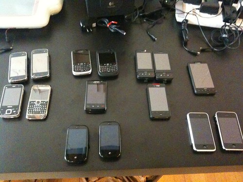 A dozen phones on a desk, including a Nokia N97 (S60v5), Blackberry Curve 8900, HTC Dream, Palm Pre, Apple iPhone 1st Gen, and Apple iPhone 3GS.